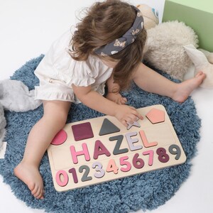 May include: A wooden puzzle with the name "HAZEL" and numbers 0-9 in various shades of pink, purple, and gray. The puzzle also includes geometric shapes. The puzzle is on a blue rug.