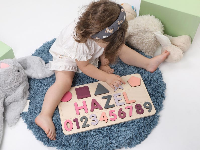 May include: A wooden puzzle with the name "HAZEL" and numbers 0-9 in various shades of pink, purple, and gray. The puzzle also includes geometric shapes. The puzzle is on a blue rug.