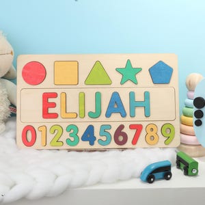 May include: Wooden puzzle with the name "ELIJAH" and numbers 0-9. The puzzle includes colorful shapes and letters. A plush lamb and toy train are in the background.
