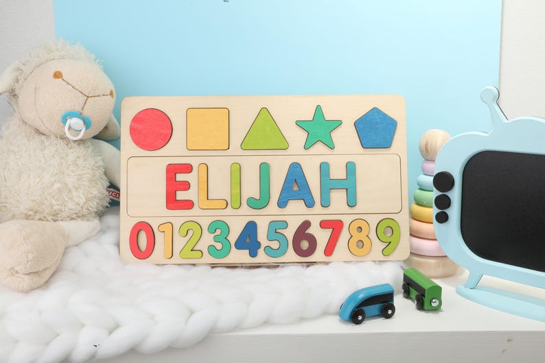 May include: Wooden puzzle with the name "ELIJAH" and numbers 0-9. The puzzle includes colorful shapes and letters. A plush lamb and toy train are in the background.