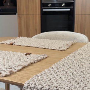 Könnte beinhalten: Beige Makramee-Tischläufer und Platzsets auf einem Holztisch. Der Läufer und die Platzsets haben ein strukturiertes, gewebtes Design mit Fransen. Ein Holzknopfdetail ist auf den Platzsets sichtbar. Küchendekoration.