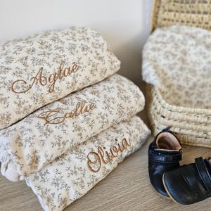 K&ouml;nnte beinhalten: Drei gefaltete cremefarbene Decken mit einem kleinen braunen Blumenmuster, jede mit einem Namen in brauner Schreibschrift bestickt: Aglae, Callie und Olivia. Ein Paar schwarze Lederschuhe f&uuml;r Babys und ein geflochtener Korb sind zu sehen.
