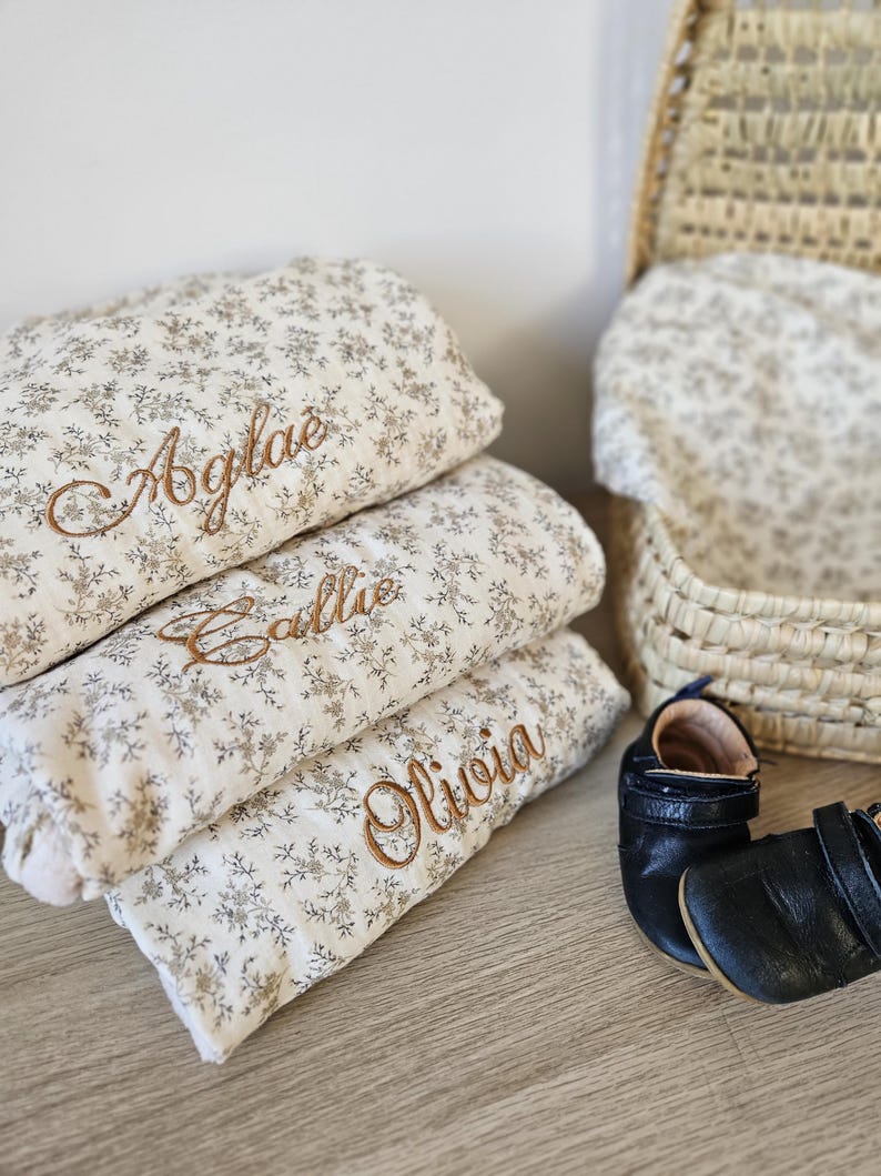 K&ouml;nnte beinhalten: Drei gefaltete cremefarbene Decken mit einem kleinen braunen Blumenmuster, jede mit einem Namen in brauner Schreibschrift bestickt: Aglae, Callie und Olivia. Ein Paar schwarze Lederschuhe f&uuml;r Babys und ein geflochtener Korb sind zu sehen.