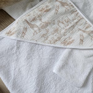 Op de afbeelding: Witte babyhanddoek met capuchon en een safari dierenprint op de capuchon. De print toont giraffen, olifanten en bomen in bruin- en beigetinten. De handdoek is gemaakt van zacht, absorberend materiaal.
