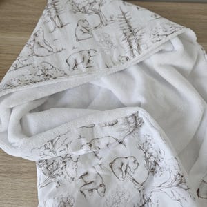 Op de afbeelding: Witte babybadhanddoek met capuchon en bruine beer- en bosprint. De handdoek is gemaakt van zacht, absorberend materiaal en heeft een capuchon met een gedetailleerd beer- en boomontwerp.