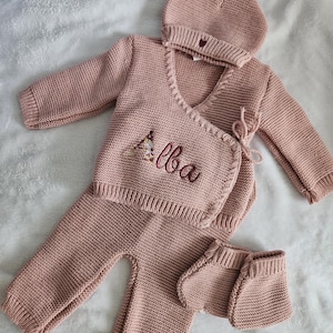 Op de afbeelding: Een oudroze gebreide babyset bestaande uit een vest met lange mouwen en overslag met de geborduurde naam Alba, een bijpassende broek, een mutsje met een klein hartje en een paar slofjes, allemaal met een gestructureerd breipatroon.