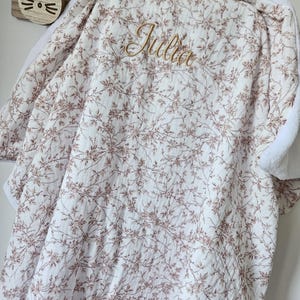 K&ouml;nnte beinhalten: Eine wei&szlig;e Babydecke mit einem zarten braunen Blumenrankenmuster und dem in Goldfaden gestickten Namen Julia. Die Decke hat auf der R&uuml;ckseite einen weichen, wei&szlig;en, strukturierten Stoff und h&auml;ngt &uuml;ber einem h&ouml;lzernen Wandhaken in Hasenform.