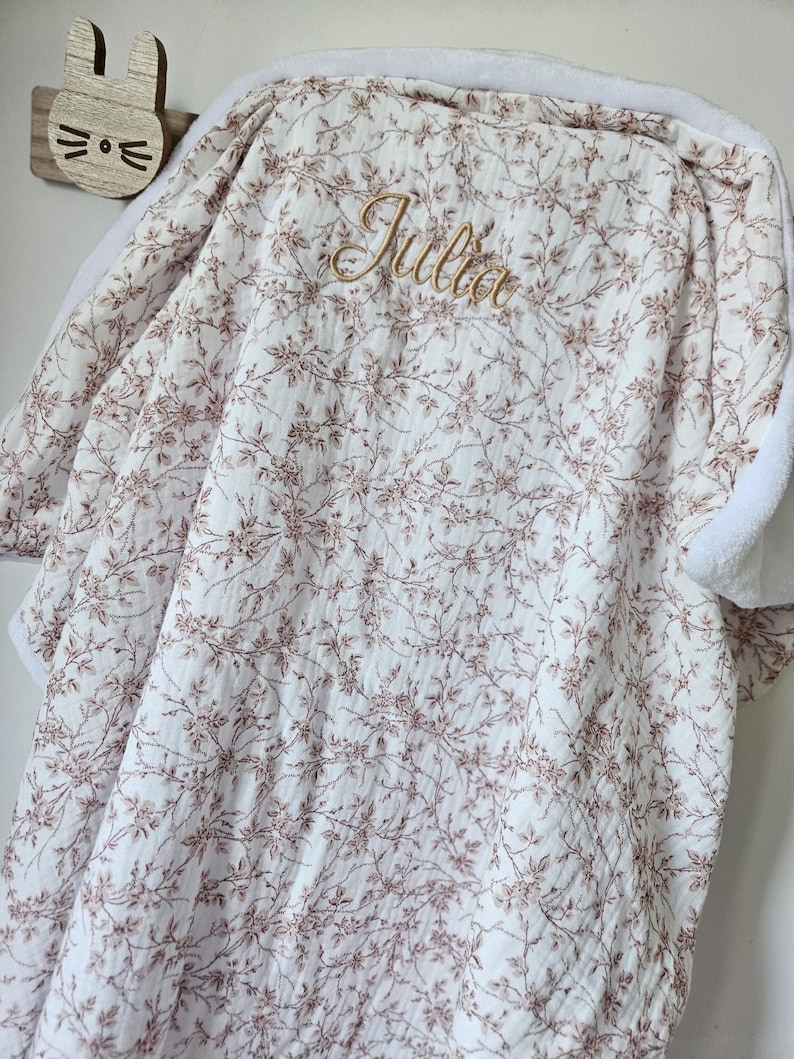 K&ouml;nnte beinhalten: Eine wei&szlig;e Babydecke mit einem zarten braunen Blumenrankenmuster und dem in Goldfaden gestickten Namen Julia. Die Decke hat auf der R&uuml;ckseite einen weichen, wei&szlig;en, strukturierten Stoff und h&auml;ngt &uuml;ber einem h&ouml;lzernen Wandhaken in Hasenform.