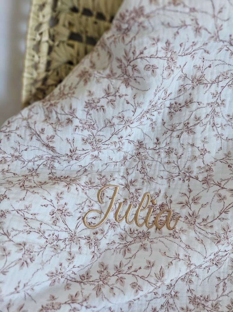 K&ouml;nnte beinhalten: Eine wei&szlig;e Musselindecke mit einem zarten braunen Blumenmuster aus kleinen Bl&auml;ttern und Bl&uuml;ten. Der Name Julia ist in goldfarbener Schreibschrift in die Mitte des Stoffes gestickt, der eine weiche, geknitterte Textur aufweist.