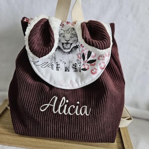 Op de afbeelding: Bordeauxkleurige corduroy rugzak met een witte flap met een sneeuwluipaard illustratie en roze bloemaccenten. De rugzak heeft een crèmekleurige handgreep en de naam "Alicia" in het wit geborduurd.