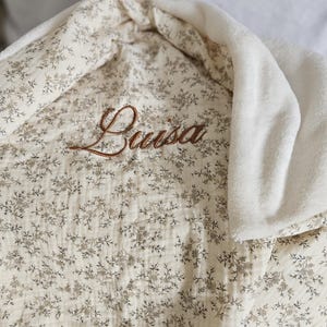 K&ouml;nnte beinhalten: Eine cremefarbene Decke mit einem zarten braunen Blumenmuster und einem weichen wei&szlig;en Fleecefutter. Der Name Luisa ist in brauner Schreibschrift in der Mitte des Stoffes eingestickt, der eine texturierte, geknitterte Optik aufweist.