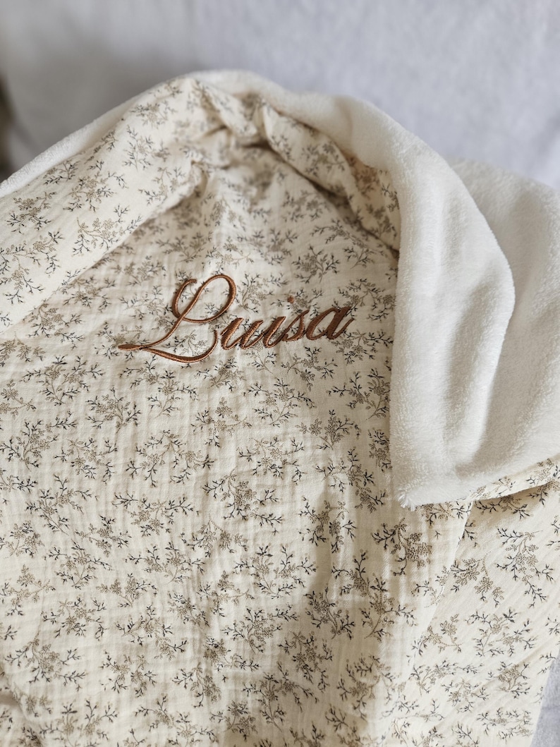 K&ouml;nnte beinhalten: Eine cremefarbene Decke mit einem zarten braunen Blumenmuster und einem weichen wei&szlig;en Fleecefutter. Der Name Luisa ist in brauner Schreibschrift in der Mitte des Stoffes eingestickt, der eine texturierte, geknitterte Optik aufweist.