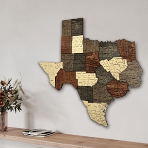 Pode incluir: Um mapa de madeira do estado do Texas com várias tonalidades de castanho, bege e cinzento. O mapa é dividido em condados, cada um rotulado com uma fonte pequena. O mapa é montado numa parede branca.