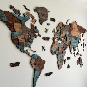 May include: A wooden world map wall art with a 3D design. The map features countries in shades of brown and blue, with text labels for oceans and continents. Includes small airplane and compass accents.