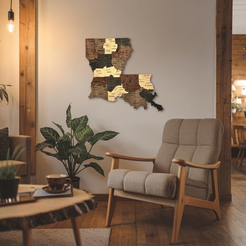 May include: A wooden map of Louisiana, featuring individual parish cutouts in varying shades of brown and tan. The map is mounted on a neutral wall, with a mid-century modern chair and coffee table in the foreground. The room has a warm, inviting atmosphere.