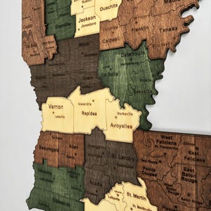 May include: A wooden map of Louisiana, USA, with each parish outlined and labeled. The map features various wood tones, including brown, green, and tan, with parish names in a simple font. The map is mounted on a white background.