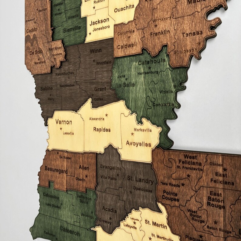 May include: A wooden map of Louisiana, USA, with each parish outlined and labeled. The map features various wood tones, including brown, green, and tan, with parish names in a simple font. The map is mounted on a white background.
