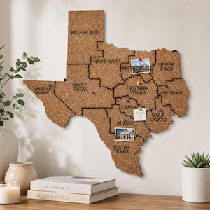 May include: A corkboard shaped like the state of Texas, divided into regions with black lines and labeled. It features push pins and small photos. The board is mounted on a white wall, with books and a candle on a wooden surface below.