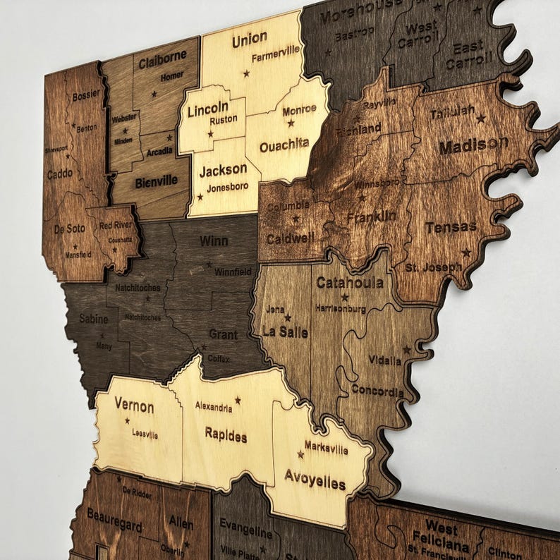 May include: A wooden map of Louisiana, USA, with each parish cut out and labeled. The map features varying shades of brown and tan wood, with parish names and cities engraved. A decorative piece for home or office.