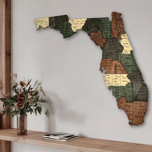 May include: A wooden map of Florida, showcasing each county with distinct colors and labels. The map is mounted on a neutral wall, with a shelf and floral arrangement in the foreground. The map's wooden texture and color variations add visual interest.