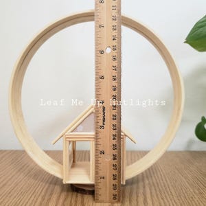 May include: A wooden miniature house with a white and pink patterned roof sits inside a wooden hoop. The hoop is 12 inches in diameter. The text "Leaf Me Up Hutlights" is visible in the image.