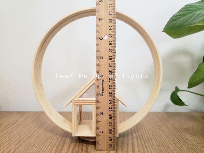 May include: A wooden miniature house with a white and pink patterned roof sits inside a wooden hoop. The hoop is 12 inches in diameter. The text "Leaf Me Up Hutlights" is visible in the image.
