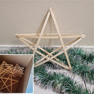 May include: A large, handmade wooden star decoration, constructed from thin wooden strips, hangs against a neutral wall. A box of similar stars sits below. Green and silver tinsel garland adds a festive touch.