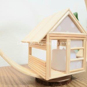 May include: A small wooden house with a white roof and a window. The house is made of popsicle sticks and is sitting on a wooden base. The text "Leaf Me Up Hutlights" is visible on the front of the house.