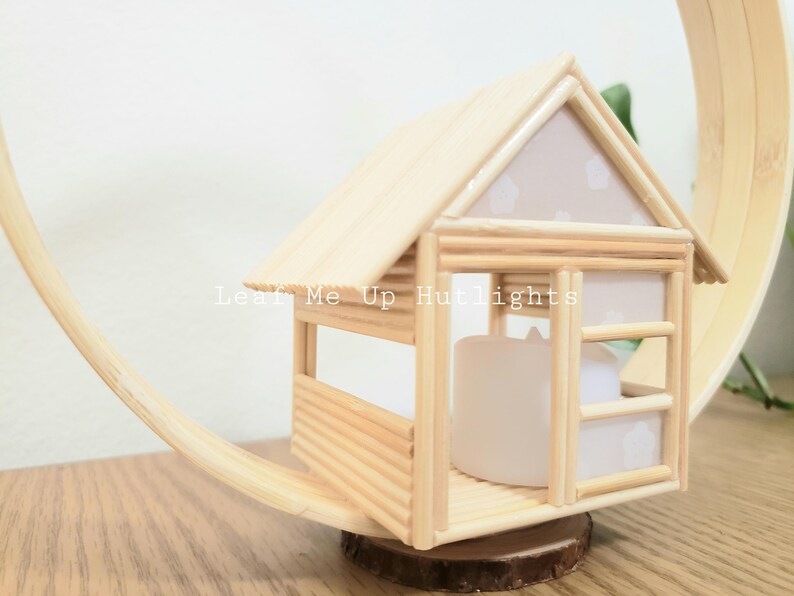 May include: A small wooden house with a white roof and a window. The house is made of popsicle sticks and is sitting on a wooden base. The text "Leaf Me Up Hutlights" is visible on the front of the house.