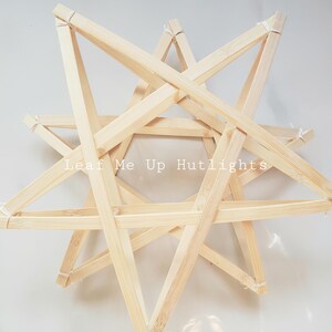 May include: A natural wood star-shaped decorative object. The star is constructed from thin wooden beams, joined at the points with white string. The text "Leaf Me Up Hutlights" is printed across the center of the star.
