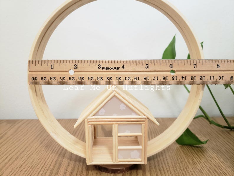 May include: A small wooden house with a white roof and a window. The house is made of popsicle sticks and is sitting on a wooden circle.