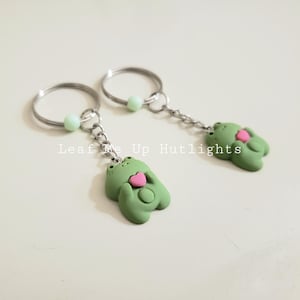 May include: Two green frog keychain charms holding pink hearts. Each charm is attached to a silver key ring with a light green bead.