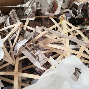 May include: A close-up of a pile of wooden frames, likely for a lamp or light fixture, covered in white paper and newspaper scraps. The text "Leaf Me Up Hutlights" is visible on the newspaper.