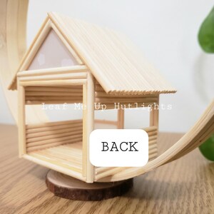 May include: A small wooden house made of popsicle sticks with a white roof and a wooden base. The house is labeled "Leaf Me Up Hutlights" and "BACK".