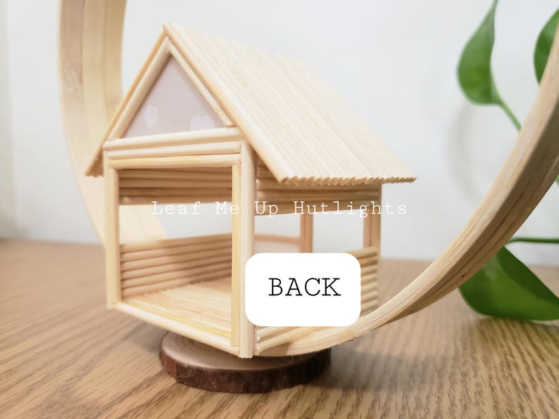 May include: A small wooden house made of popsicle sticks with a white roof and a wooden base. The house is labeled "Leaf Me Up Hutlights" and "BACK".