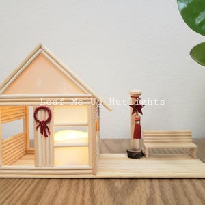May include: A miniature wooden house with a glowing interior, a small red wreath, and a decorative star. The house is made of popsicle sticks and has a small bench and a decorative post next to it. The text "Leaf Me Up Hut Lights" is visible.