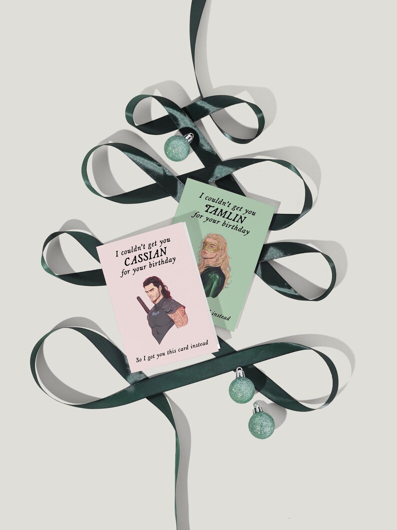May include: Two greeting cards with illustrations of characters from a fantasy novel. The card on the left features a man with a sword and the text "I couldn't get you Cassian for your birthday. So I got you this card instead." The card on the right features a woman with long blonde hair and the text "I couldn't get you Tamlin for your birthday. So I got you this card instead."