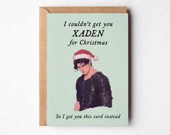 May include: A Christmas card with a light green background and a brown envelope. The card features an illustration of a person wearing a Santa hat and black armor. Text on the card reads, "I couldn't get you XADEN for Christmas. So I got you this card instead."