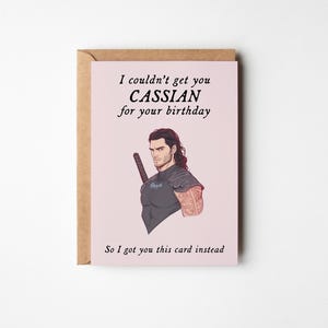 May include: A pink birthday card featuring a cartoon illustration of a man with brown hair and a sword. The card reads "I couldn't get you Cassian for your birthday. So I got you this card instead."