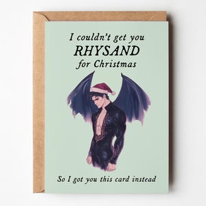 May include: A greeting card with a light green background and a brown envelope. The card features an illustration of a man with dark wings and a Santa hat. Text on the card reads, "I couldn't get you RHYSAND for Christmas. So I got you this card instead."