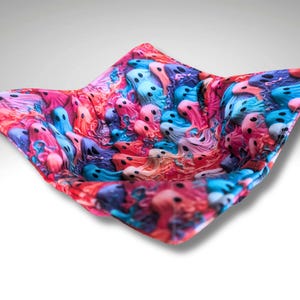 May include: A square fabric bowl with a colourful ghost pattern. The bowl features a repeating design of small, cartoon ghosts in shades of pink, blue, and purple. The fabric is folded to create a bowl shape.