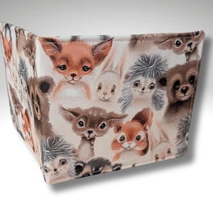 May include: A beige fabric wallet with a repeating pattern of cute animal faces. The animals include a fox, a deer, a squirrel, a hedgehog, and a rabbit. The animals are all drawn in a cartoon style with brown, white, and gray colors.