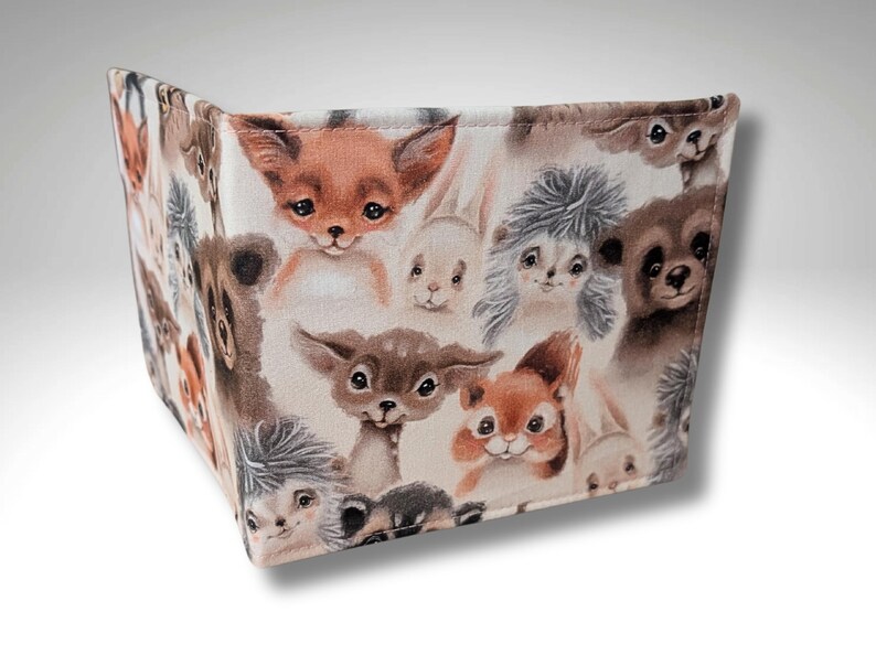 May include: A beige fabric wallet with a repeating pattern of cute animal faces. The animals include a fox, a deer, a squirrel, a hedgehog, and a rabbit. The animals are all drawn in a cartoon style with brown, white, and gray colors.