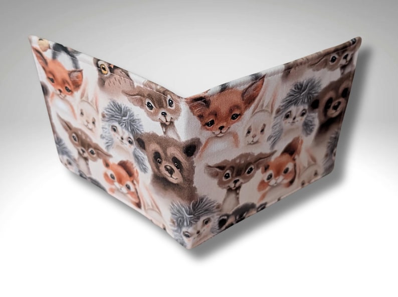 May include: A white fabric wallet with a repeating pattern of cute animal faces. The animals include foxes, deer, squirrels, rabbits, and owls. The wallet is folded closed.