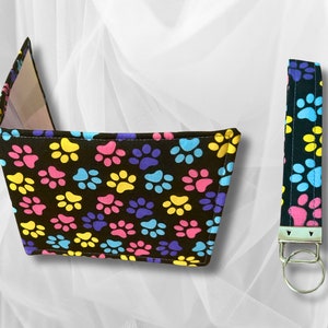 May include: A black fabric key chain with a colourful paw print pattern and a silver key ring. The key chain is attached to a black fabric wallet with a colourful paw print pattern.