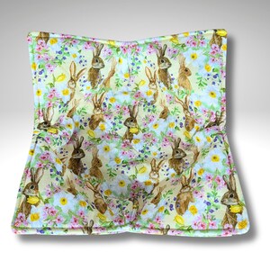 May include: A square, quilted bowl cosy with a repeating pattern of watercolour bunnies and flowers. The fabric has a light yellow background with pink, blue, and yellow floral accents. The bunnies are brown and white.
