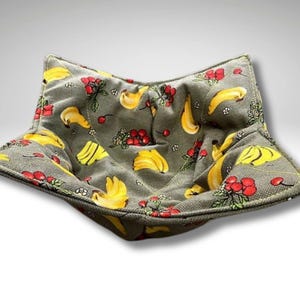 May include: A green fabric bread basket with a banana and red berry pattern. The basket is shaped like a star and has a soft, quilted texture.