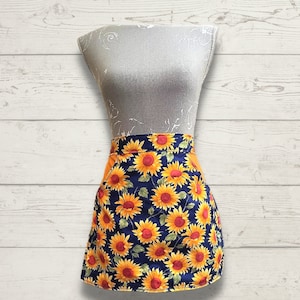 May include: A blue and yellow sunflower print apron with a solid orange waistband. The apron is on a white wooden background.
