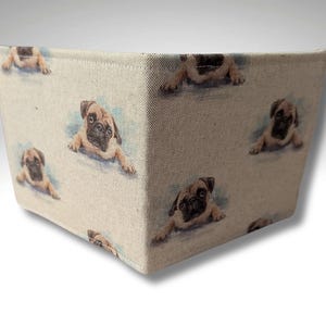 May include: A beige fabric storage bin featuring a repeating pattern of pug dog illustrations. The pugs are light brown with black facial features and are lying down. The bin has a rectangular shape.