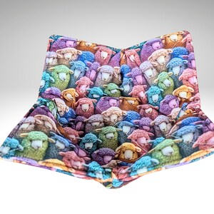 May include: A bowl cosy with a repeating pattern of cartoon sheep in shades of blue, green, purple, and orange. The sheep have white faces. The cosy is open and ready to hold a bowl, ideal for keeping food warm.